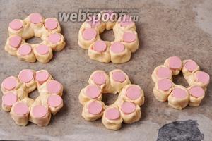 Завернуть каждую сосиску (350 грамм) по кругу, выложив кусочки сосисок срезом вгору (вниз) в виде цветка и выложив на противень, застеленный пергаментом. Если вы не уверены в качестве пергамента, можно смазать его небольшим количеством подсолнечного масла (3 мл). 1 сосиску в тесте разрезать на кусочки до конца и по одному кусочку с сосиской выложить в центр цветка. Накрыть булочки пищевой плёнкой и оставить в тёплом месте на 30 минут.