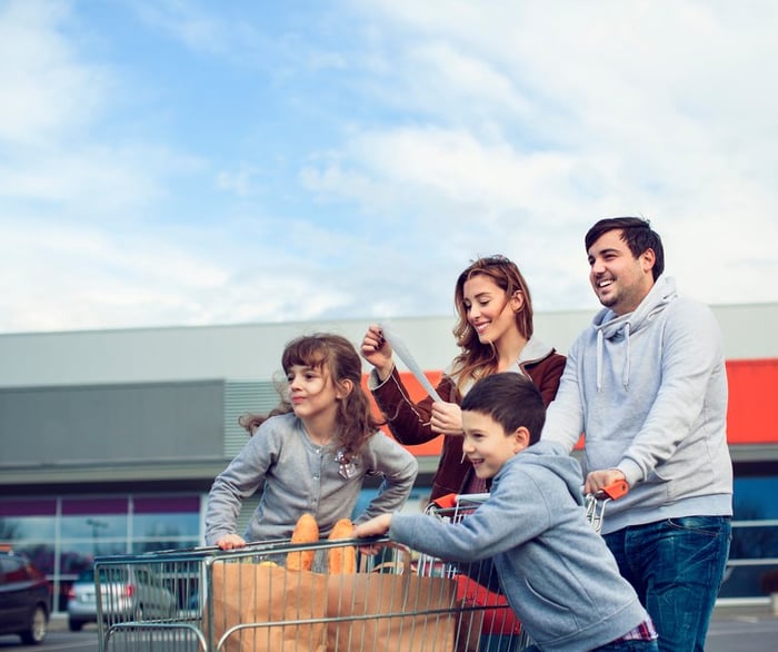 Two parents and two kids pushing a shopping cart full of groceries while checking the receipt