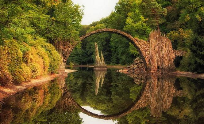 Мост Ракотцбрюке – место, которые нужно увидеть хоть раз в своей жизни. Глядя на него, сложно освободится от мистического ощущения. Не зря его также называют Дьявольским. Почему? Необычный мост находится в старинном дендропарке Кромлау на берегу озера Ракоц в Германии. Его разбил в 1844 году Фридман Герман Рётшке. Это примерно 120 километров от Дрездена. Парк огромный, в нем есть озера и пешеходные тропы. Здесь можно увидеть скульптуры, изображающие Геркулеса и разных богов, а также оказаться в рае и в аду, которые воплотились в виде двух гротов: один из них светлый, а другой – темный. Парк хорошо оборудован: у дороги есть парковка, а в нескольких километрах – кемпинг, доступный по стоимости. Обычно людей здесь не очень много, но порой приезжают целые автобусы туристов.  Это изумительное сооружение. Мост создан из базальтовых колонн в 1860 году. Базальт здесь не находили, поэтому его везли из Швейцарии и Богемии. Из него создали также скалы, гроты и пирамиды, которые украшают парк. Мост представляет собой идеальное в своей полуокружности сооружение. Именно его удивительная геометрия связала с ним множество мифов и поверий. Это 35-метровая каменная дуга, перекинутая над водой. Над всем этим нависают ветви раскидистых деревьев. Независимо от того, где вы стоите, он и его отражение будут сливаться в идеальный круг. Важная роль в этом отводится уровню воды. Однако легенды объясняют это участием в строительстве моста самого нечистого. Серый и загадочный материал, из которого создана переправа, базальтовые зубья, вырывающиеся из земли перед входом на мост, безмятежная водная гладь, тишина и умиротворение природы создают здесь уникальную атмосферу. В этом месте невозможно не ощутить благоговейный трепет. Невозможно представить даже, что все это создано человеческими руками. Кто создал Чертов мост? Имя того, кто создал Дьявольский мост, неизвестно, но с ним связаны первые легенды. Говорят, архитектор, которому поручили строительство, не отличался талантом, но мечтал прославиться. Неуверенный в своих силах, он пошел на заключение сделки с дьяволом, согласившимся помочь в обмен на душу первого живого существа, которое ступит на мост. Нечистый вселился в архитектора, создал удивительное творение. После этого он покинул тело и начал ждать обещанную плату. Архитектор выпустил на мост пса. Раздосадованный черт прыгнул в воду.  Какие бы поверья ни ходили, но архитектор сотворил действительно уникальную достопримечательность. Как смотрятся острые базальтовые пластины, которые выступают из воды и суши! Они обрамляют мост, зловеще отражаются в воде. Кроме того, гениальный создатель моста расположил его в крайне удачном месте. Озеро Ракоц обладает идеальным уровнем воды, нужным для создания визуального эффекта идеального круга. А конструкция поддерживает его таким дольше, чем это бывает в естественных условиях. Современные легенды Говорят, если в полнолуние сможешь проплыть под мостом в лодке так, чтобы не увидеть ни разу отражение и не коснуться воды, то ты обретешь сверхспособности, а еще исполнятся все желания. Есть представление о том, что мост – это портал, благодаря которому можно оказаться в другом мире. Эти слухи подогревает то, что в 1983 году здесь исчезли два туриста из Швеции. Штрафы смельчакам Мост притягивает туристов, но последним ходить по нему строго запрещено. За это полагается административный штраф. Сделано это для того, чтобы как можно дольше сохранить этот уникальный памятник в целости и сохранности. Вокруг входов путь туристам преграждают заборы и специальные вывески. Фотографии людей, идущих по мосту и прыгающих на нем либо фотошоп, либо нарушение закона. Хоть и нельзя фотографироваться на мосту, это можно сделать в других уголках парка, территория которого достигает 200 гектаров. Самое подходящее время посещения – август и сентябрь. В это время здесь особенно красиво. Золотая листва создает особую атмосферу на озере. Очень хорошо здесь и в мае, когда цветут рододендроны и азалии. Как приехать Вход в парк бесплатный, а открыт он круглый год. Проще всего добраться сюда из Дрездена или Берлина на машине или поезде. Правда, если вы поедете поездом, придется делать пересадки. Например, из Берлина нужно доехать до Котбуса, пересесть на поезд до Вайсвассера, а там – на автобус до парка Кромлау. Есть еще один интересный способ полюбоваться Дьявольским мостом – воспользоваться узкоколейкой. Она сама – достопримечательность. К счастью, ее маршрут проходит через нужный парк. Вагончики отправляются каждые выходные. Станция отправления – Weibwasser. Мистические места можно найти еще много где в Германии. Например, пользуются популярностью каменные гроты Экстернштейне, замок Вольфзегг с привидением, гора Лорелей, «Сонная деревня» и многое другое. Среди всего этого – ни на что не похожий Дьявольский мост.