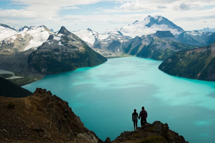 Two hikers holding hands and standing on a lookout point over a glacial lake surrounded by snow-capped mountains.