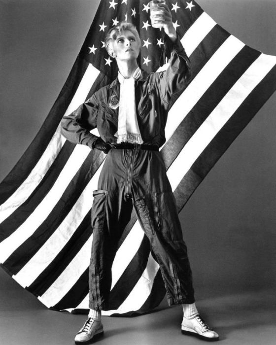 Монохромный портретный снимок британского рок-музыканта и певца в комбинезоне пилота на фоне американского флага.