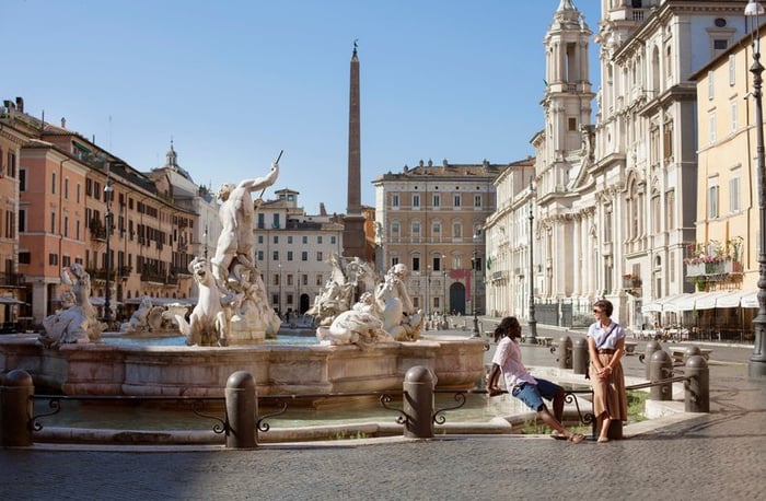 Two vacationers sitting next to a beautiful fountain in an open square in Rome, Italy.
