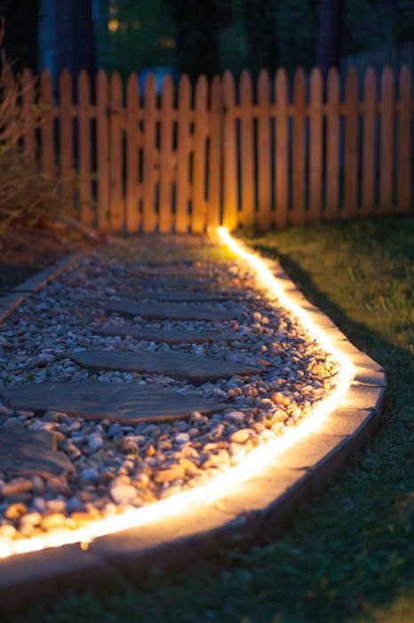 Садовая дорожка с неоновой подсветкой, которая создана из деревянных досок на фоне белой гальки. 