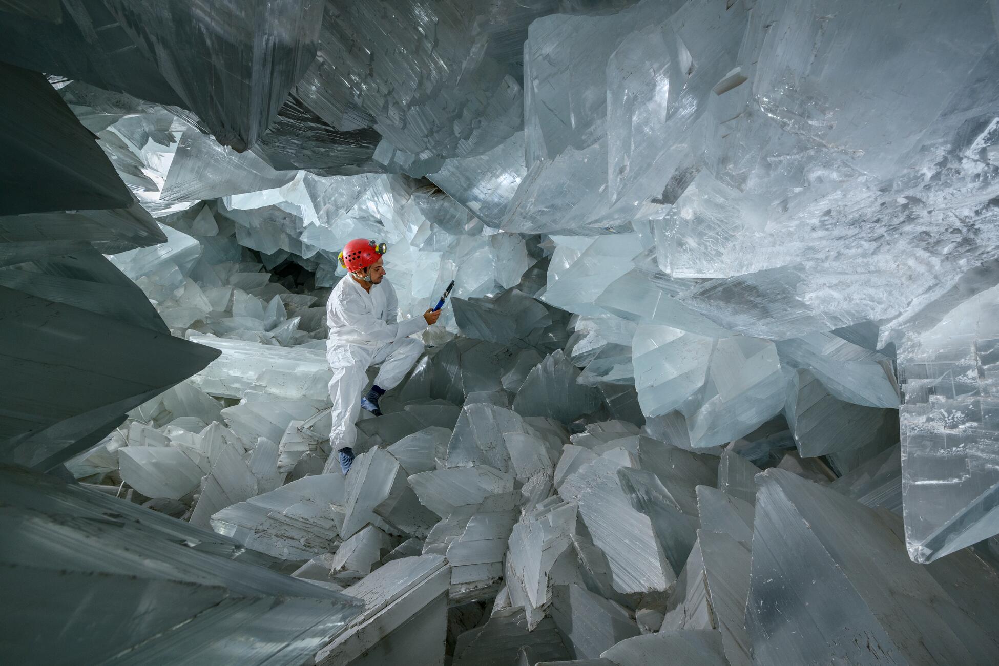 Picture of a man in a white protective suit and red hardhat holds a device in his hand and sits in a large cave full of huge white and gray crystals on all surfaces