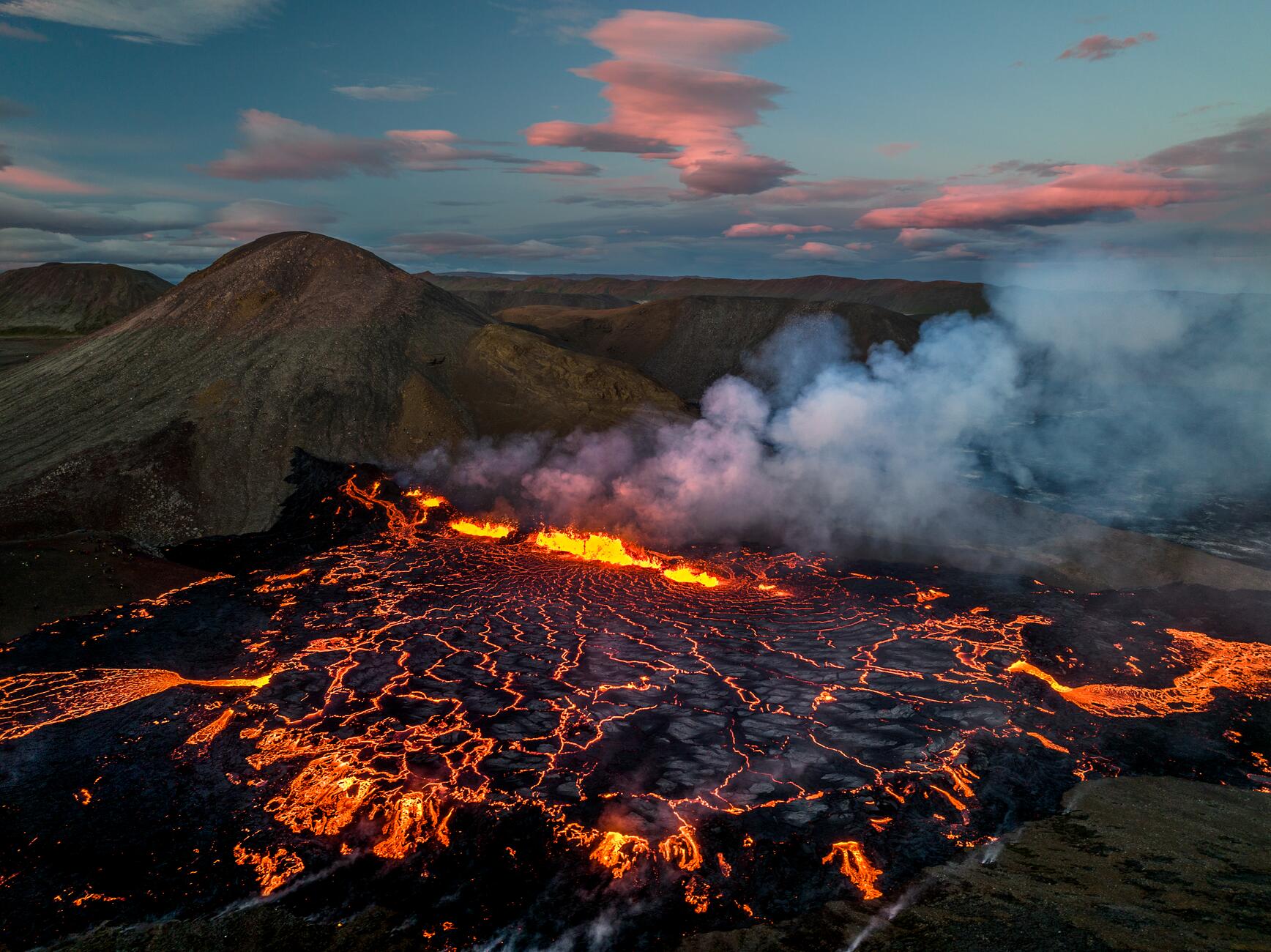 Фотография вулкана в Исландии с большим бассейном темной лавы с несколькими ярко-оранжевыми извержениями лавы, происходящими в бассейне