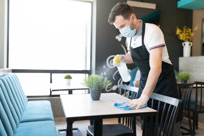 A waiter wearing a medical mask and cleaning a table in a restaurant with a spray bottle and cloth.