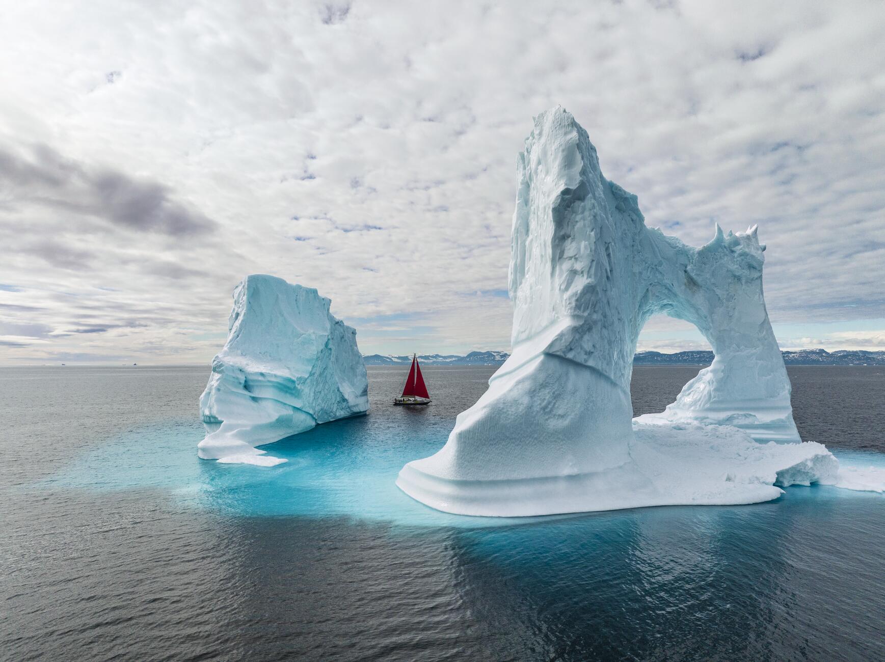 Фотография парусника с красными парусами, который плывет между двумя очень большими айсбергами. Один айсберг напоминает арку с разрывом посередине