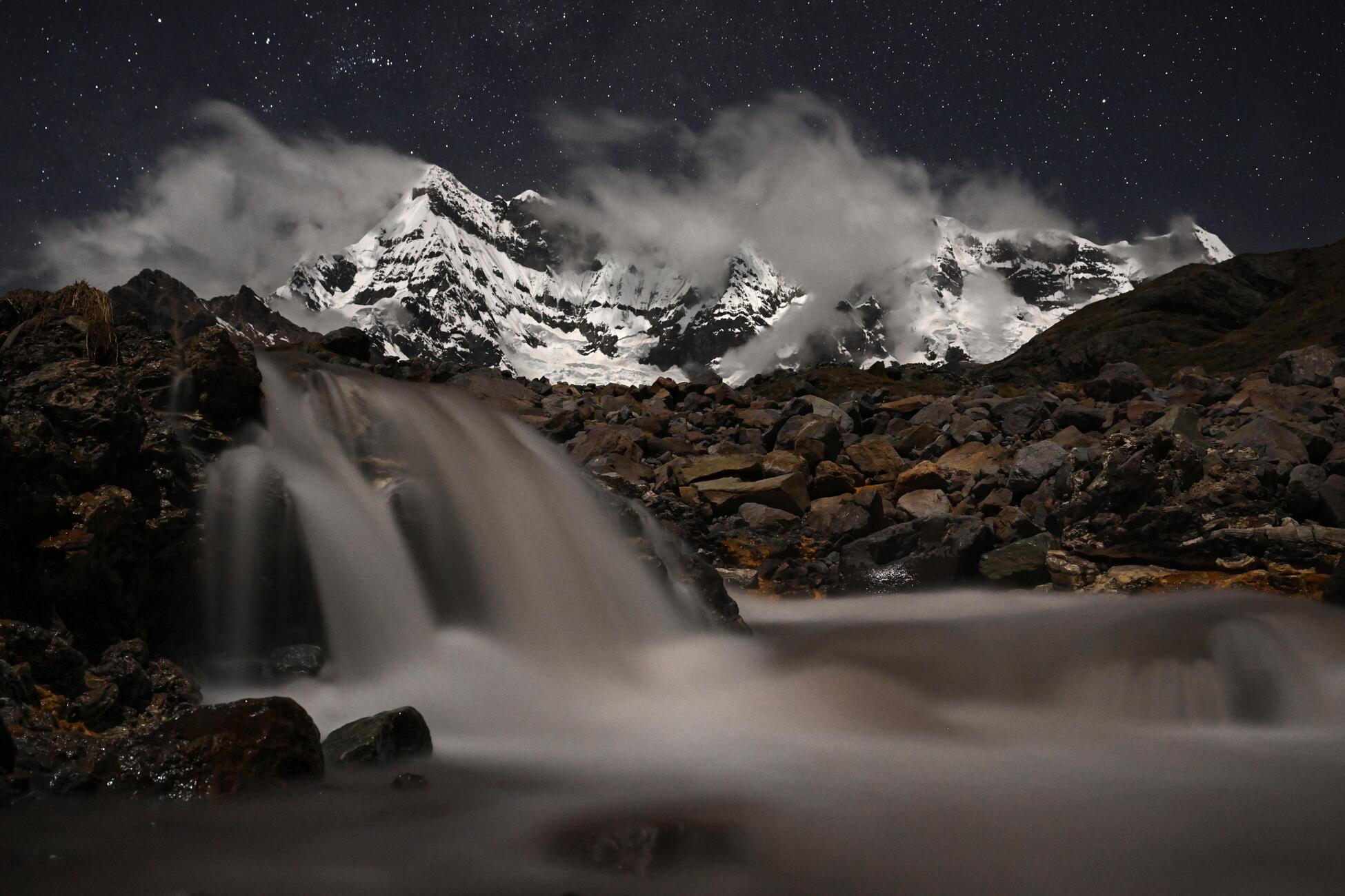 Photograph of a waterfall at night with large snow covered mountains in the background
