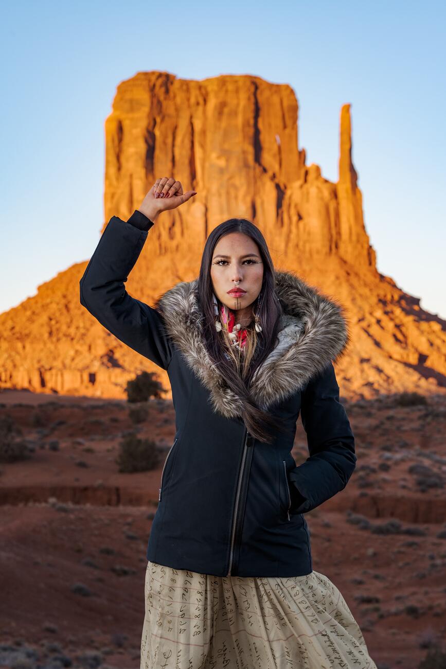 Picture of a woman with tattooed lines on her chin and fingers. Stands in front of a large natural brown sandstone mountain with vertical walls. She is wearing a dark coat with a fur lined hood and a tan dress while raising one hand in the air with her fist closed