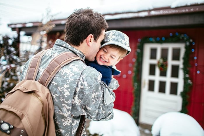 Military father with backpack hugging child outside home.