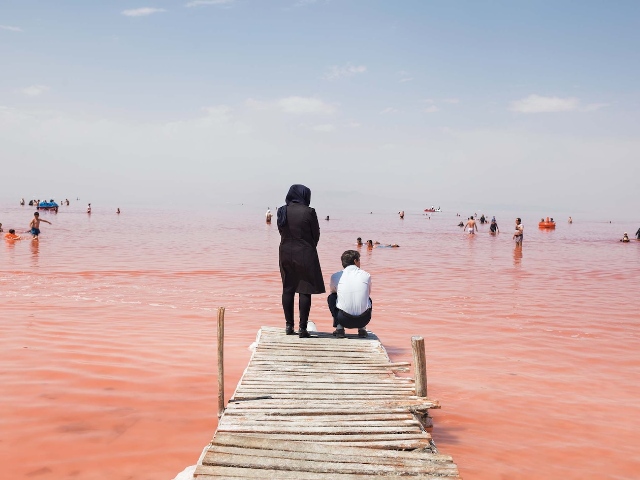 Женщина стоит, а мужчина сидит на корточках на причале над красновато-розовым озером