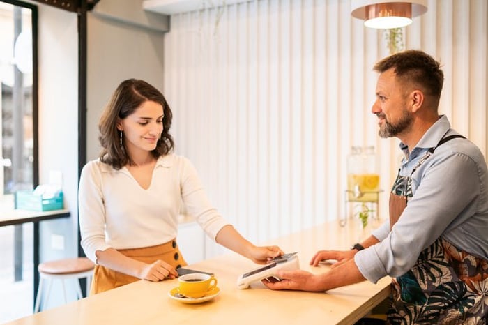 A woman tapping her credit card on a payment reader while standing across the counter from a barista.