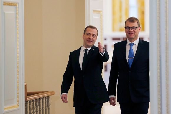 Venäjän pääministeri Dmitri Medvedev ja Suomen pääministeri Juha Sipilä tulevat suurista ovista. Miehillä on yllään mustat puvut, Sipilällä on sininen kravatti ja Medvedevillä tumma. Molemmat hymyilevät. Medvedev viittoo huoneen sisälle päin.