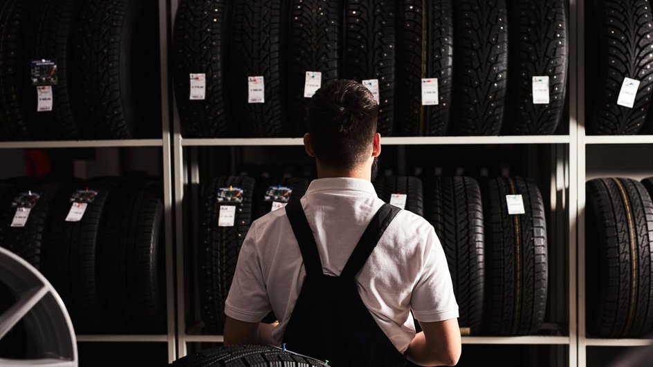 Вид сзади на молодого механика-мужчину в униформе, разглядывающего автомобильные шины в автосервисе.