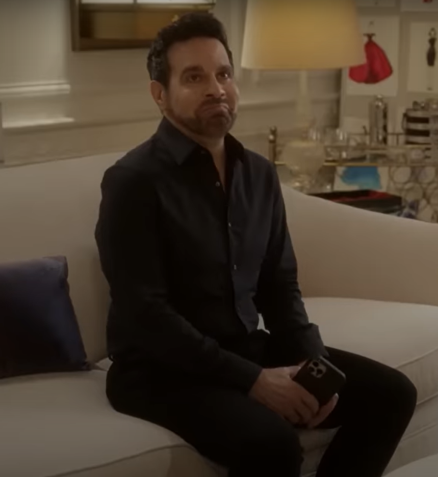 Mario Cantone as Anthony Marentino sits on a white sofa wearing a black shirt and black pants