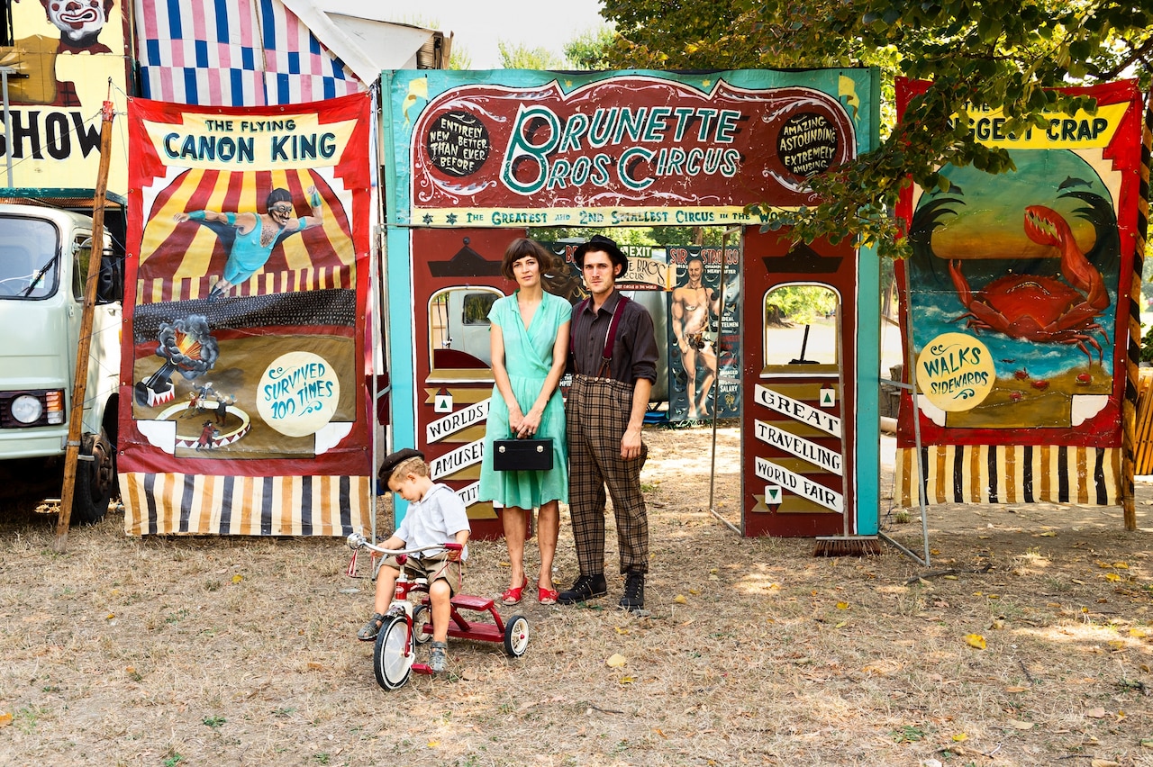 Фотография пары и их сына на трехколесном велосипеде, позирующих для семейного портрета перед входом в цирк.