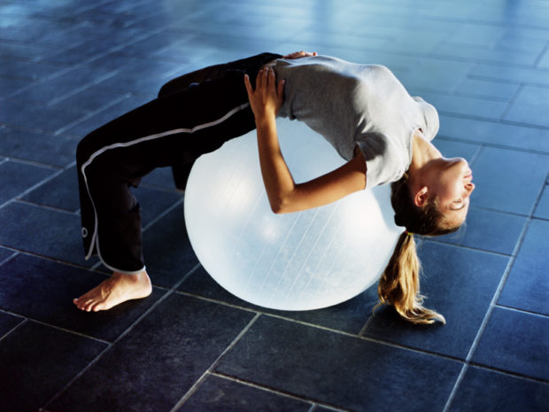 Девушка с волосами в хвостике лежит на белом полупрозрачном фитболе и смотрит вдаль