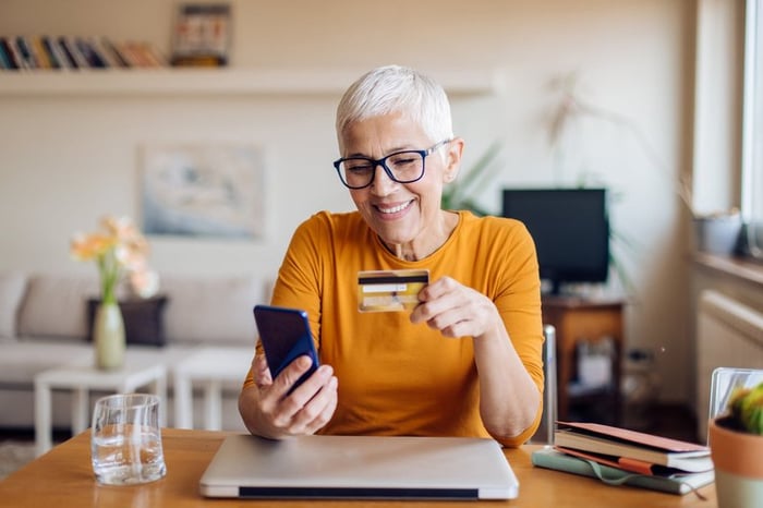 A mature woman smiles while sitting at her table. She is holding a cellphone and a credit card.
